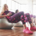Zwei schwangere Frauen in Sportkleidung sitzen auf Stabilitätsbällen in einer hellen Turnhalle, lächeln und trainieren zusammen. Beide tragen rosa Sportkleidung und Turnschuhe.