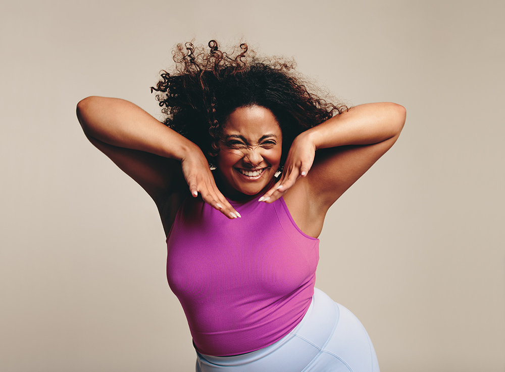 Eine lächelnde Frau mit lockigem Haar posiert spielerisch und hält ihre Hände unter ihr Kinn. Für das Beckenbodentraining trägt sie ein leuchtend lila ärmelloses Oberteil und eine hellblaue Hose vor einem neutralen Hintergrund.