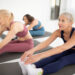 Drei ältere Frauen in Sportkleidung sitzen in einem hellen Raum auf Yogamatten, strecken sich nach vorne und berühren ihre Zehen, um an einem Gruppenfitness- oder Yogakurs teilzunehmen.