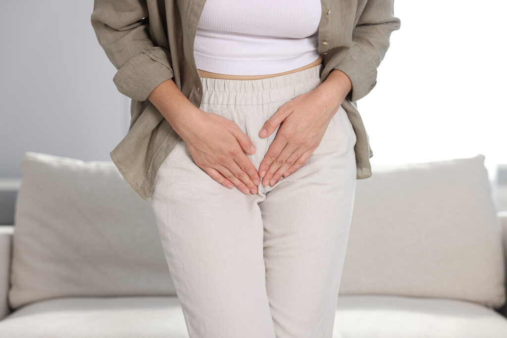 Eine Frau, die in einem Haus steht und beide Hände auf ihren Unterleib presst, scheint sich unwohl zu fühlen oder Schmerzen zu haben. Sie trägt eine helle Hose, ein weißes Oberteil und ein offenes Hemd mit Knopfleiste.
