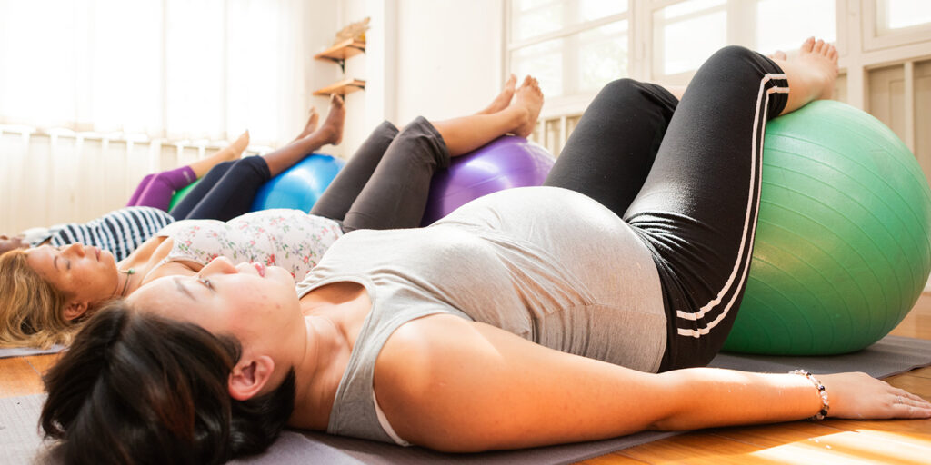 Drei schwangere Frauen liegen auf Yogamatten in einem Raum und stützen ihre Beine auf große Gymnastikbälle, um an einem pränatalen Fitnesskurs teilzunehmen. Das Sonnenlicht strömt durch die Fenster und schafft eine ruhige Atmosphäre.