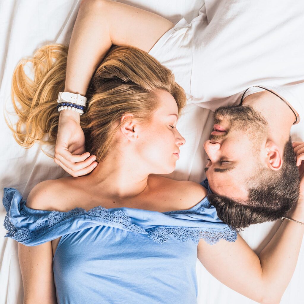 Eine Frau und ein Mann liegen sich auf einem Bett gegenüber, die Augen geschlossen, die Köpfe nah beieinander und die Arme sanft aneinandergelegt. Die Frau trägt ein blaues schulterfreies Oberteil und der Mann ein weißes Hemd.