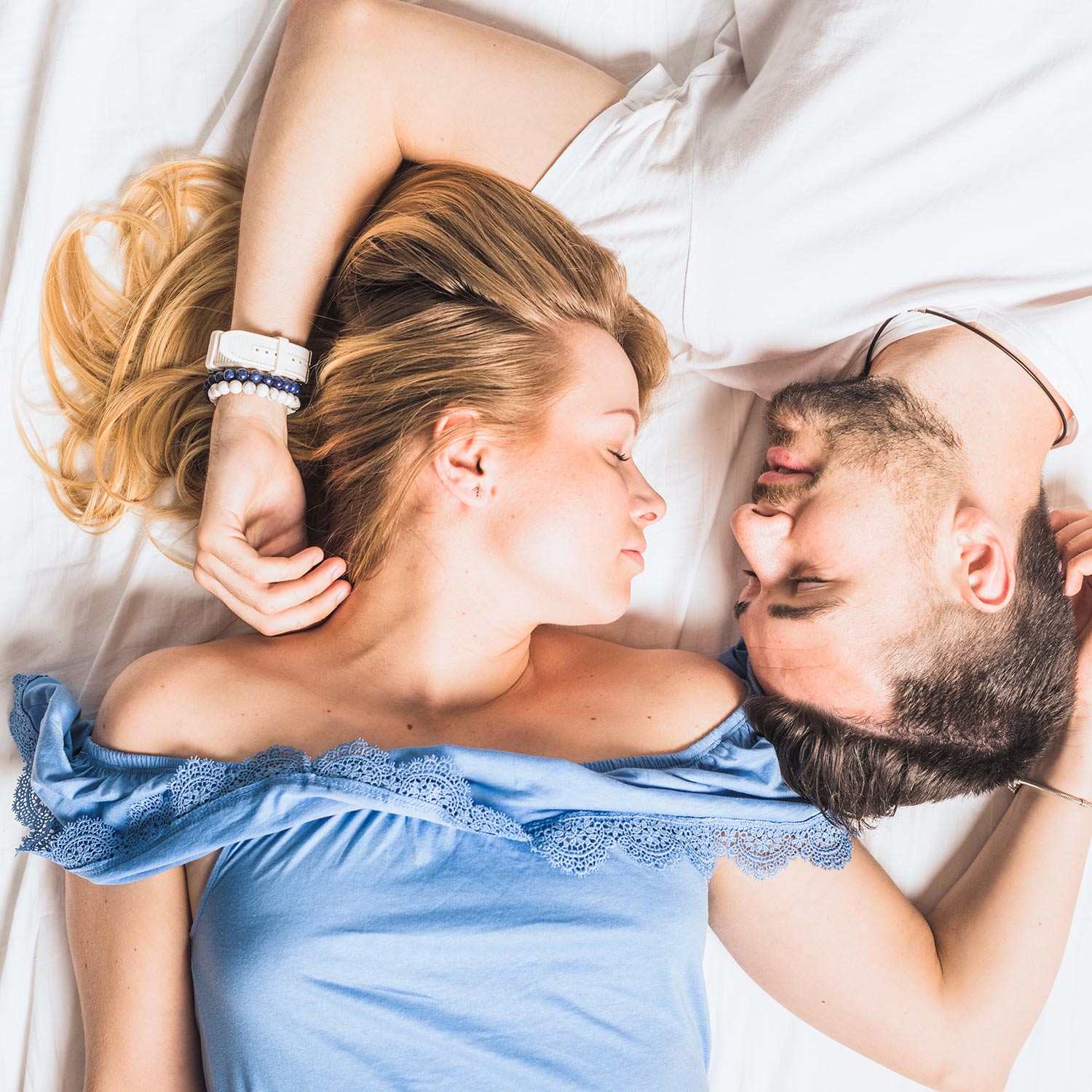 Eine Frau und ein Mann liegen sich auf einem Bett gegenüber, die Augen geschlossen, die Köpfe nah beieinander und die Arme sanft aneinandergelegt. Die Frau trägt ein blaues schulterfreies Oberteil und der Mann ein weißes Hemd.