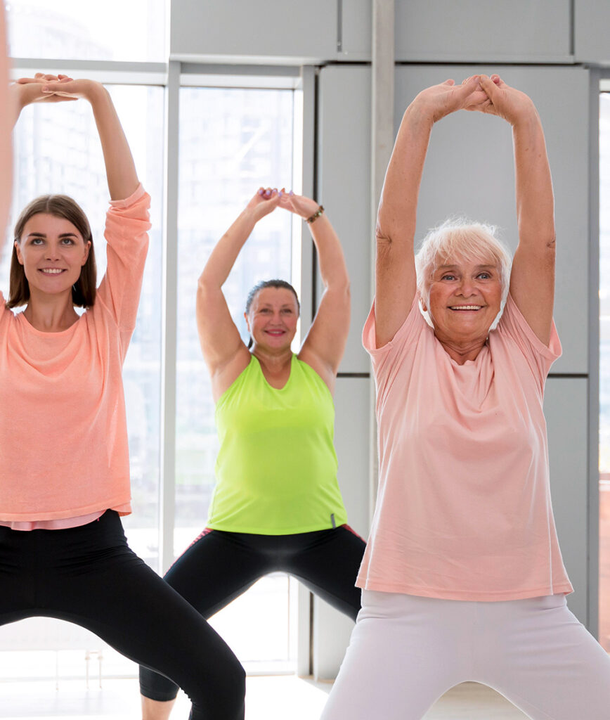 Drei Frauen unterschiedlichen Alters stehen in einem hellen Raum, lächeln und strecken sich mit über den Kopf gehobenen Armen, in bequemer Trainingskleidung.