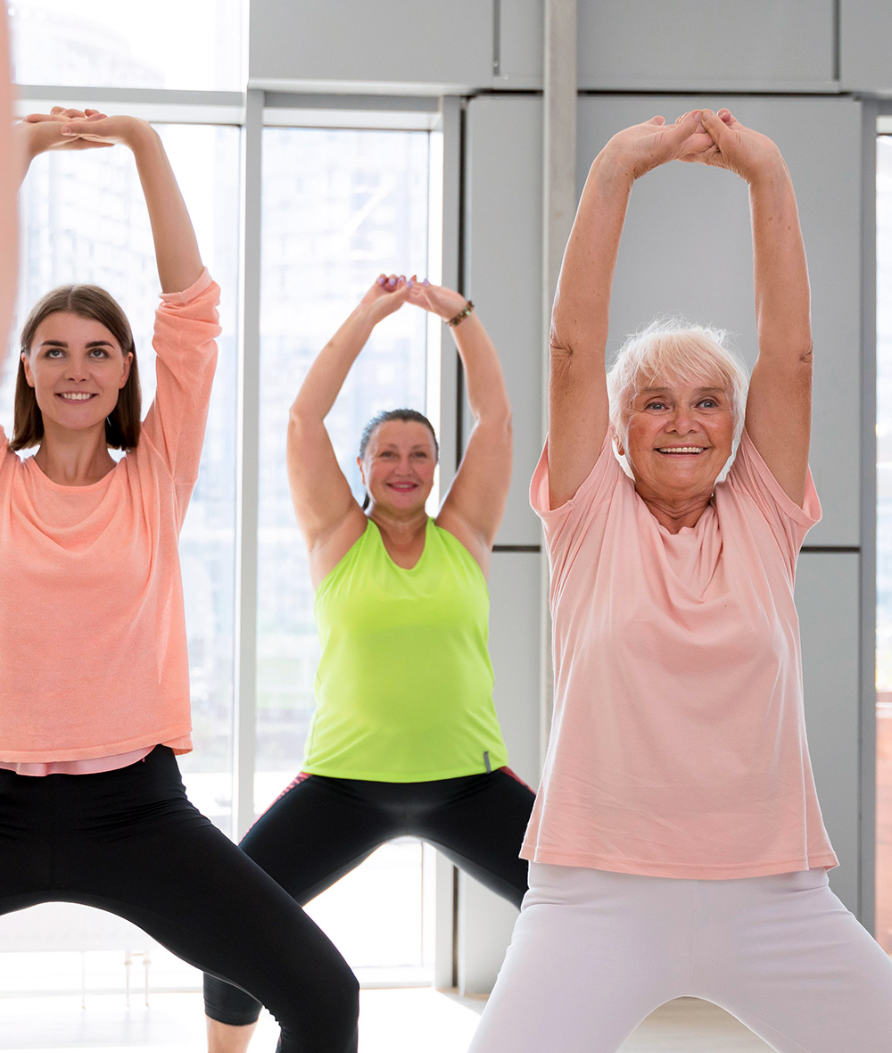 Drei Frauen unterschiedlichen Alters stehen in einem hellen Raum, lächeln und strecken sich mit über den Kopf gehobenen Armen, in bequemer Trainingskleidung.